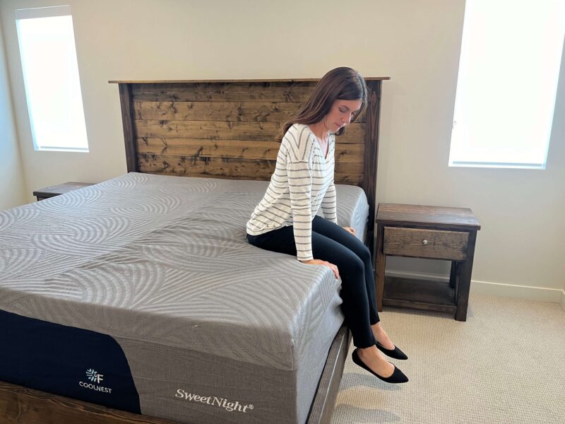 Kristina Hindes sits on the edge of the SweetNight CoolNest mattress, looking down at its surface.