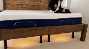 Person sitting on an Amerisleep adjustable bed holding a remote control, with warm amber under-bed lighting glowing beneath the navy blue base and wood bed frame.