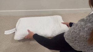 A person holding a white pillow horizontally with both hands, bending it slightly to test how well it springs back to its original shape.