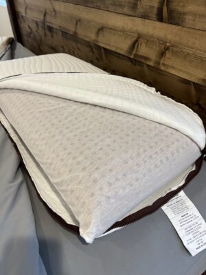 A close-up of two pillows stacked on a bed showing their fabric textures and care labels, with one pillow partially unzipped to reveal its inner cover, photographed against a wooden headboard backdrop