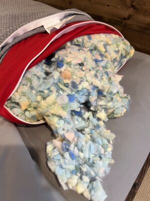 A gray and red zippered pillow unzipped and tipped over to spill out a large amount of multicolored shredded foam fill in blue, green, yellow, and white pieces.